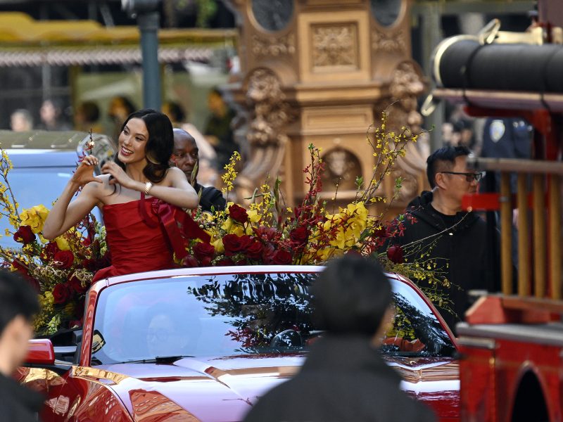 A woman in a red dress sits atop a red convertible decorated with flowers, holding a small object and smiling during a parade. People and vehicles are visible around her.