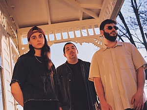 Three people stand under a decorative gazebo. One wears a backwards cap, another has short hair and a mustache, and the third has a beard and sunglasses.