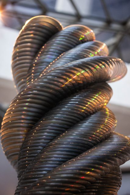 Close-up of a large, twisted metallic sculpture with reflective surfaces that catch and scatter colored light. Geometric ceiling structure visible in the background.