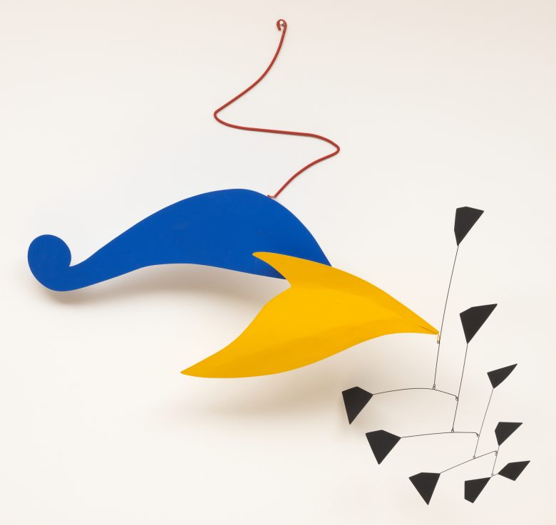 Abstract mobile sculpture with a large blue curved shape, a yellow pointed shape, a red wire, and a cluster of small black geometric forms hanging below.