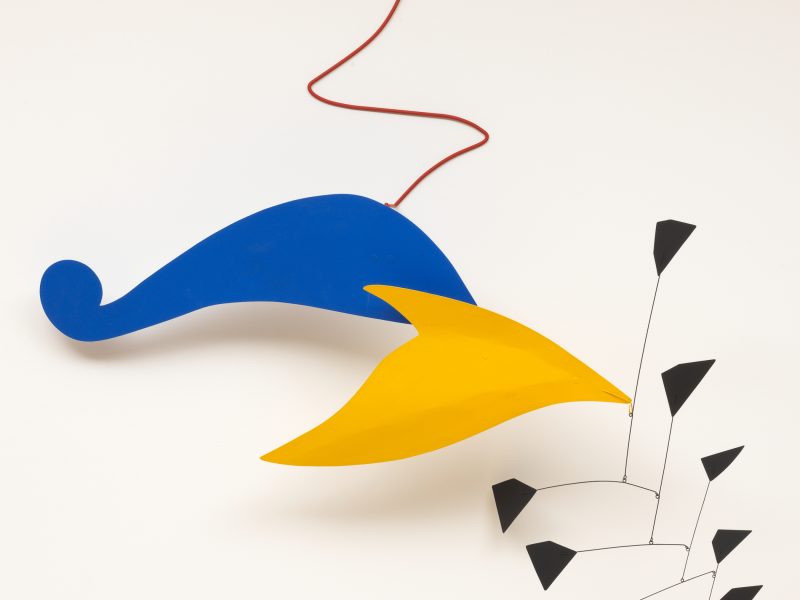 Abstract mobile sculpture with a large blue curved shape, a yellow pointed shape, a red wire, and a cluster of small black geometric forms hanging below.
