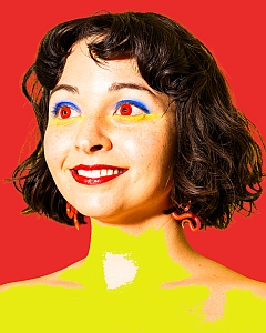 A woman with short dark hair, bright red lipstick, vivid blue and yellow eye makeup, and red earrings stands against a solid red background. Her skin and neck are edited to appear bright yellow.