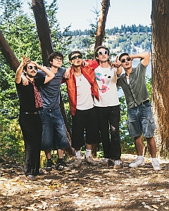 Five men stand together outdoors in a wooded area near water, posing casually with arms around each other and looking toward the camera.