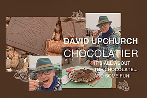 A man in glasses and a green hat enjoys eating chocolate at a market stall, with chocolate pieces and cocoa beans displayed around him. Text reads "David Upchurch Chocolatier.
