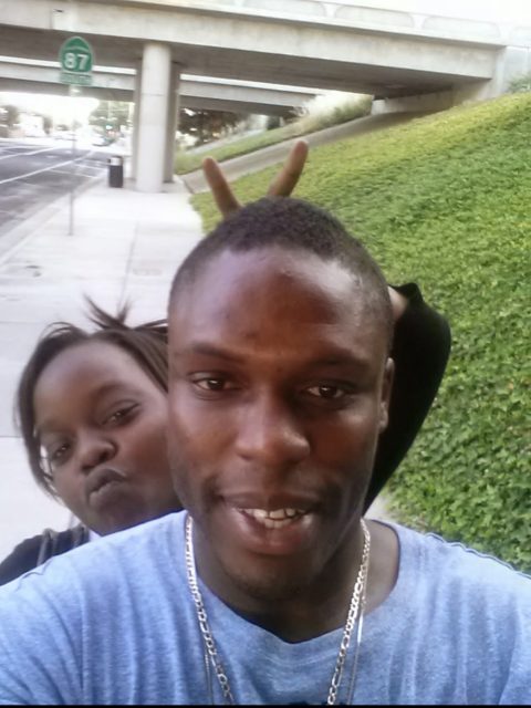 Two people pose for a selfie on a sidewalk near an overpass; the person in back makes a peace sign behind the other’s head.