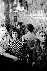 Four men stand in a tiled room with mirrored walls and a hanging light fixture, some facing forward and others with their backs to the camera.