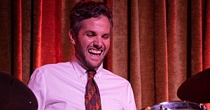 A man in a white shirt and patterned tie smiles while playing the drums in front of gold curtains.
