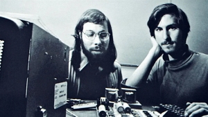 Two young men sit at a desk with early computer equipment and electronic components, looking at the camera in a black and white photo.