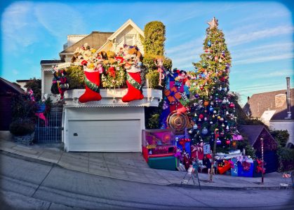 A house on a steep street is elaborately decorated for Christmas with large stockings, Santa figures, ornaments, a tall Christmas tree, and oversized gift boxes.