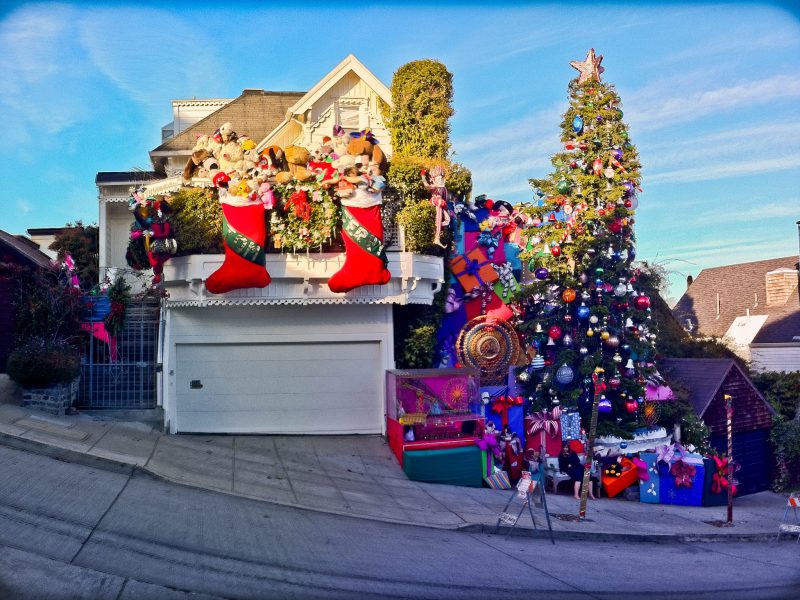 A house on a steep street is elaborately decorated for Christmas with large stockings, Santa figures, ornaments, a tall Christmas tree, and oversized gift boxes.