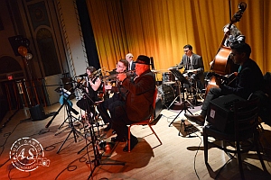 A jazz band performs on stage with brass, woodwind, piano, drums, and upright bass in front of a yellow curtain.