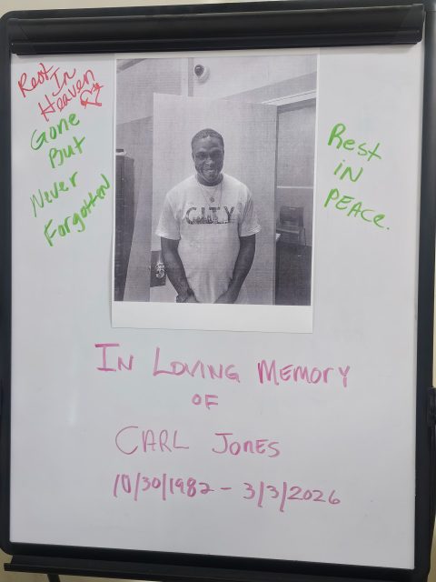 A photo of a man is taped to a whiteboard with handwritten messages reading "Rest in Heaven," "Rest in Peace," and "In Loving Memory of Carl Jones, 10/30/1982 - 3/3/2026.