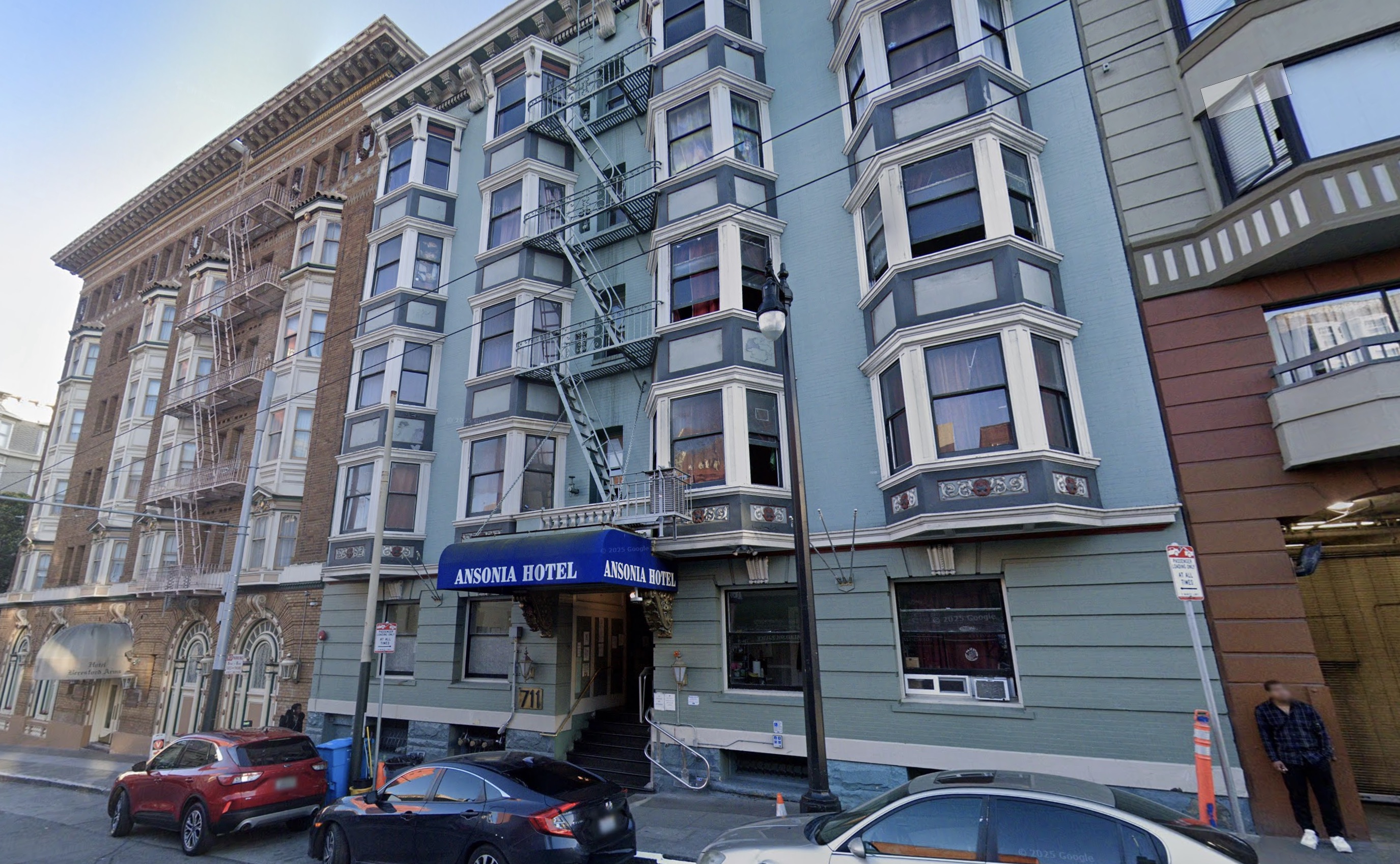 Street view of the Ansional Hotel, a multi-story building with bay windows, a blue awning, and cars parked along the curb.