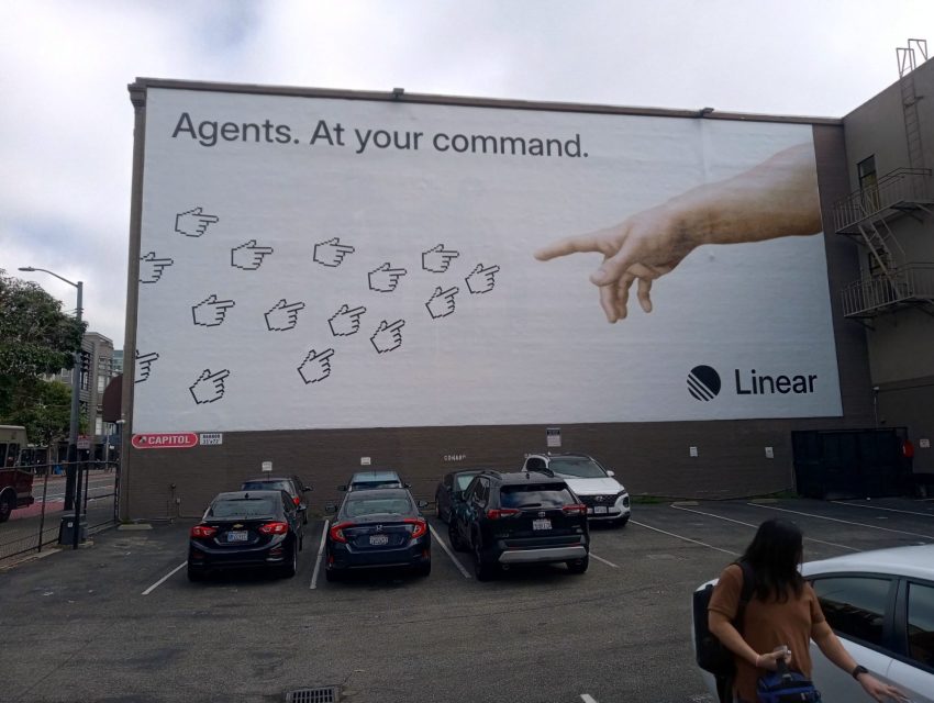 Large mural on a building shows a realistic hand commanding multiple cursor icons, with the text "Agents. At your command." and the Linear logo. Cars are parked below.