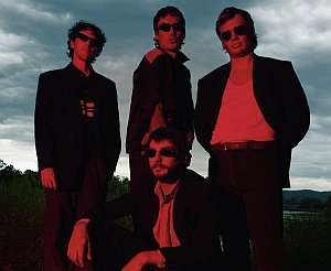 Four men wearing dark sunglasses and suits pose outdoors under a cloudy sky, with a red-tinted light illuminating them.