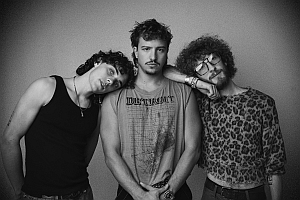 Three young men pose together against a plain background; one leans his head on another's shoulder. All wear casual clothing, including a tank top, a sleeveless shirt, and a leopard print top.
