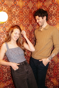 Two people stand in front of a patterned wall, smiling and laughing. The woman has long hair and is wearing a sleeveless top; the man is wearing a long-sleeve shirt and has one hand in his pocket.