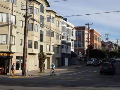 A person rides a bicycle through an intersection on a sunny city street lined with apartments, parked cars, and utility poles.