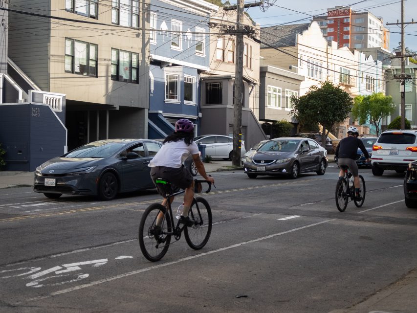 Two cyclists ride in a bike lane on a city street with cars parked and driving nearby, and residential buildings in the background.