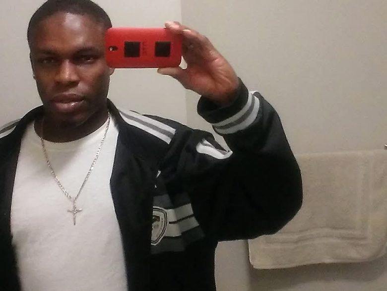 A man stands in front of a bathroom mirror taking a selfie with a red phone, wearing a black jacket, white shirt, and a cross necklace. A towel hangs on the wall behind him.