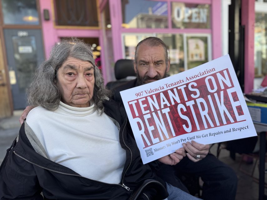 Two people sit outside a building holding a sign that reads "Tenants on Rent Strike" as part of the 907 Valencia Tenants Association.