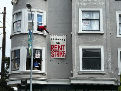 A person hangs a large banner reading "Tenants on Rent Strike" from a window of a gray apartment building while another person sits in a lower window.