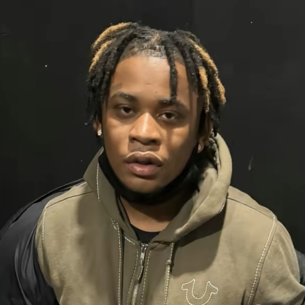 A young man with short dreadlocks and light tips wears a khaki hoodie and black face mask under his chin, sitting against a dark background.