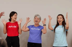 Three women standing side by side raise their hands and smile, wearing colorful T-shirts against a plain background.