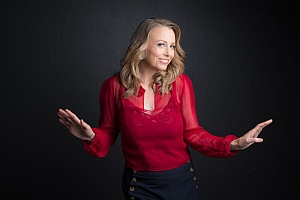 Woman with blonde hair wearing a red blouse and black pants stands against a black background, smiling and holding her hands up as if gesturing or dancing.