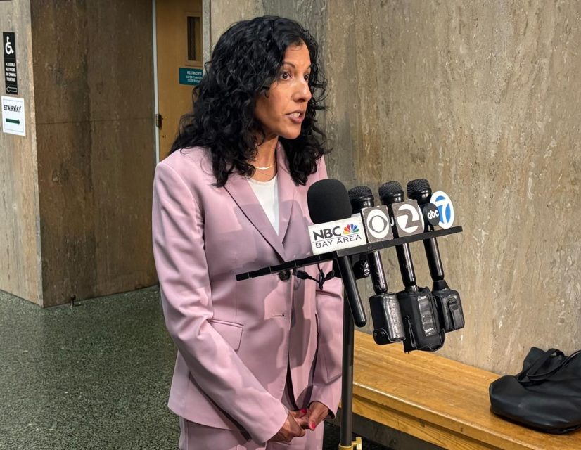 A woman in a pink suit speaks at a microphone podium with several news station logos in a hallway next to a wooden bench.