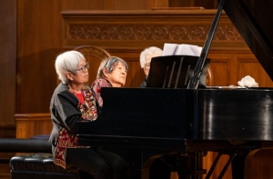 Three elderly women play a grand piano together in a wood-paneled room, focused on sheet music resting on the piano stand.