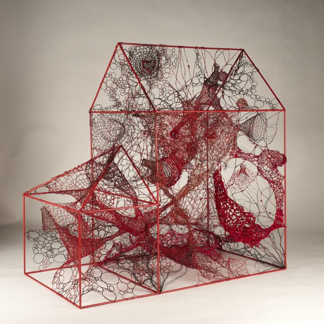 A wireframe sculpture of a house with red and black intertwined threads and circular patterns filling the interior, set against a plain background.
