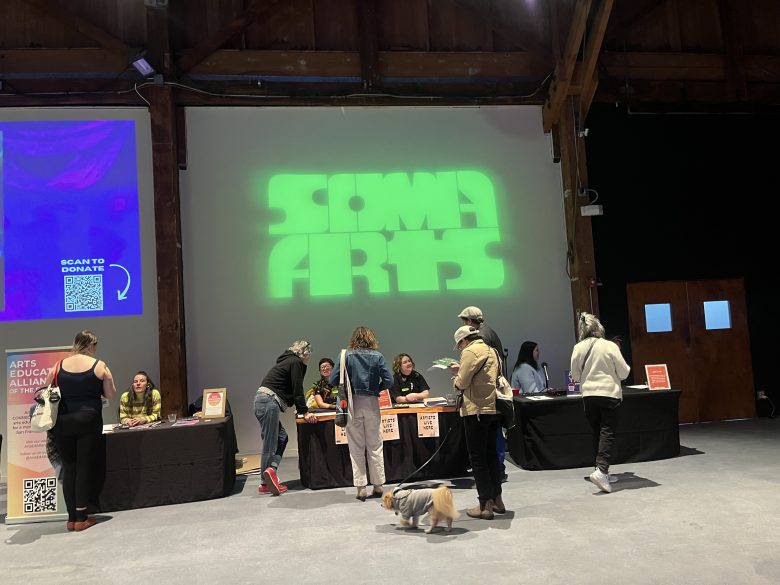 People interact at information tables under a green "SOMA ARTS" projection inside a spacious event hall. A small dog is present in the foreground.