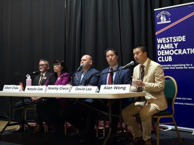 Five people sit at a table with nameplates, microphones, and water bottles during a panel event hosted by the Westside Family Democratic Club. A banner stands to the right.