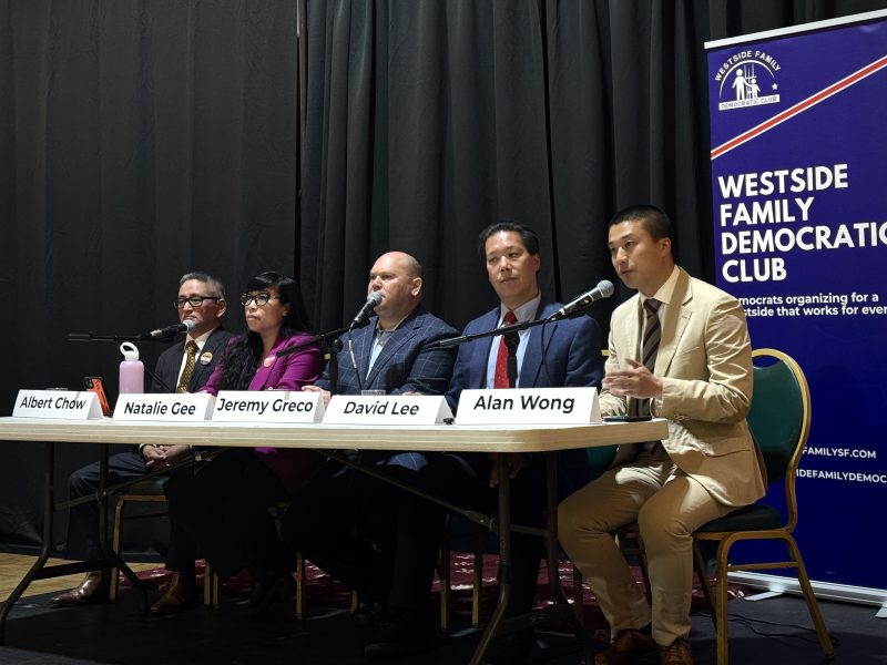 Five people sit at a table with nameplates, microphones, and water bottles during a panel event hosted by the Westside Family Democratic Club. A banner stands to the right.