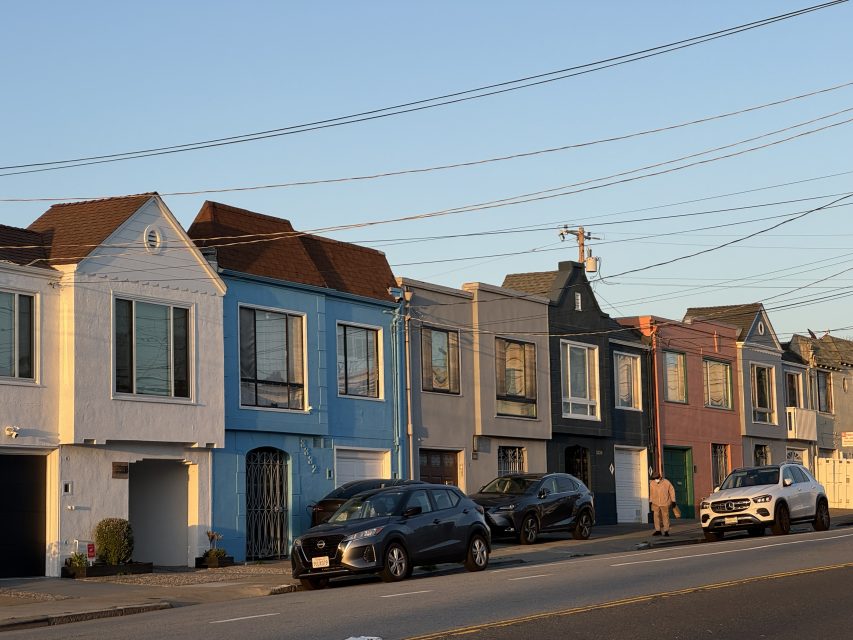 Row of colorful, two-story houses with parked cars and a person walking on the sidewalk in warm late afternoon light.