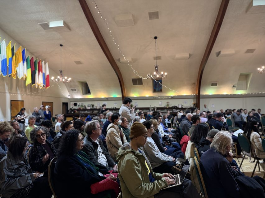 A large group of people sits in rows of chairs inside a hall with international flags, string lights, and high arched ceilings.