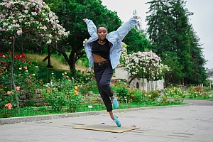 A person wearing a light blue jacket and blue shoes leaps in the air on a paved path in a park with blooming flowers and green trees in the background.