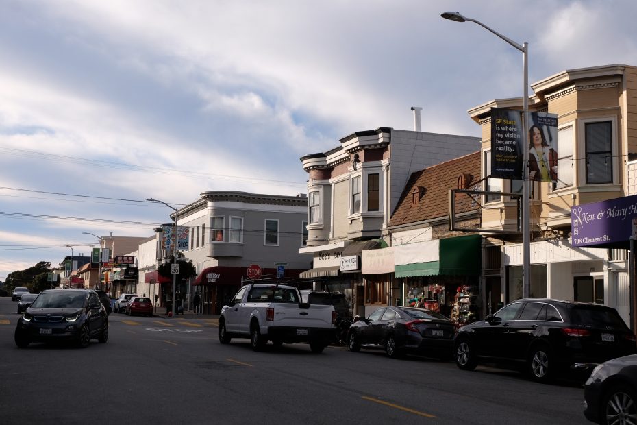 A street with parked cars, small businesses, and shops in two-story buildings under a partly cloudy sky.