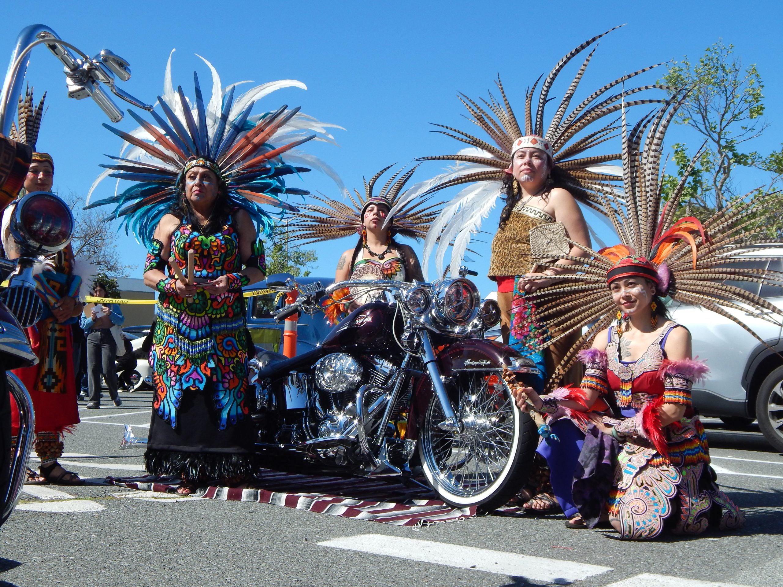 Five people wearing colorful traditional Aztec attire and feathered headdresses pose around a motorcycle in a parking lot on a sunny day.