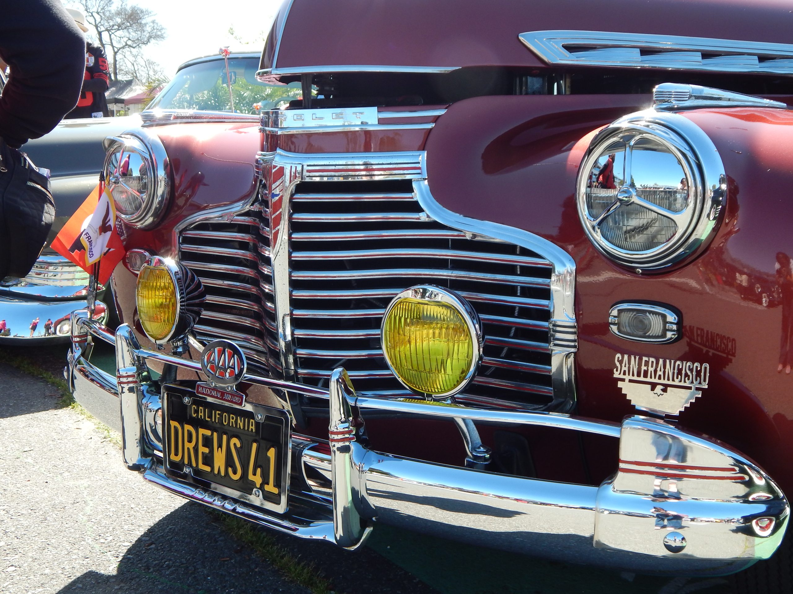 Front view of a vintage maroon car with chrome details, yellow headlamps, and a California license plate reading "DREWS 41" displayed at a car show.