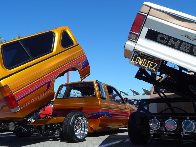 Two colorful custom trucks with lifted beds, showing painted designs and chrome wheels, parked outdoors at a car show under a clear blue sky.