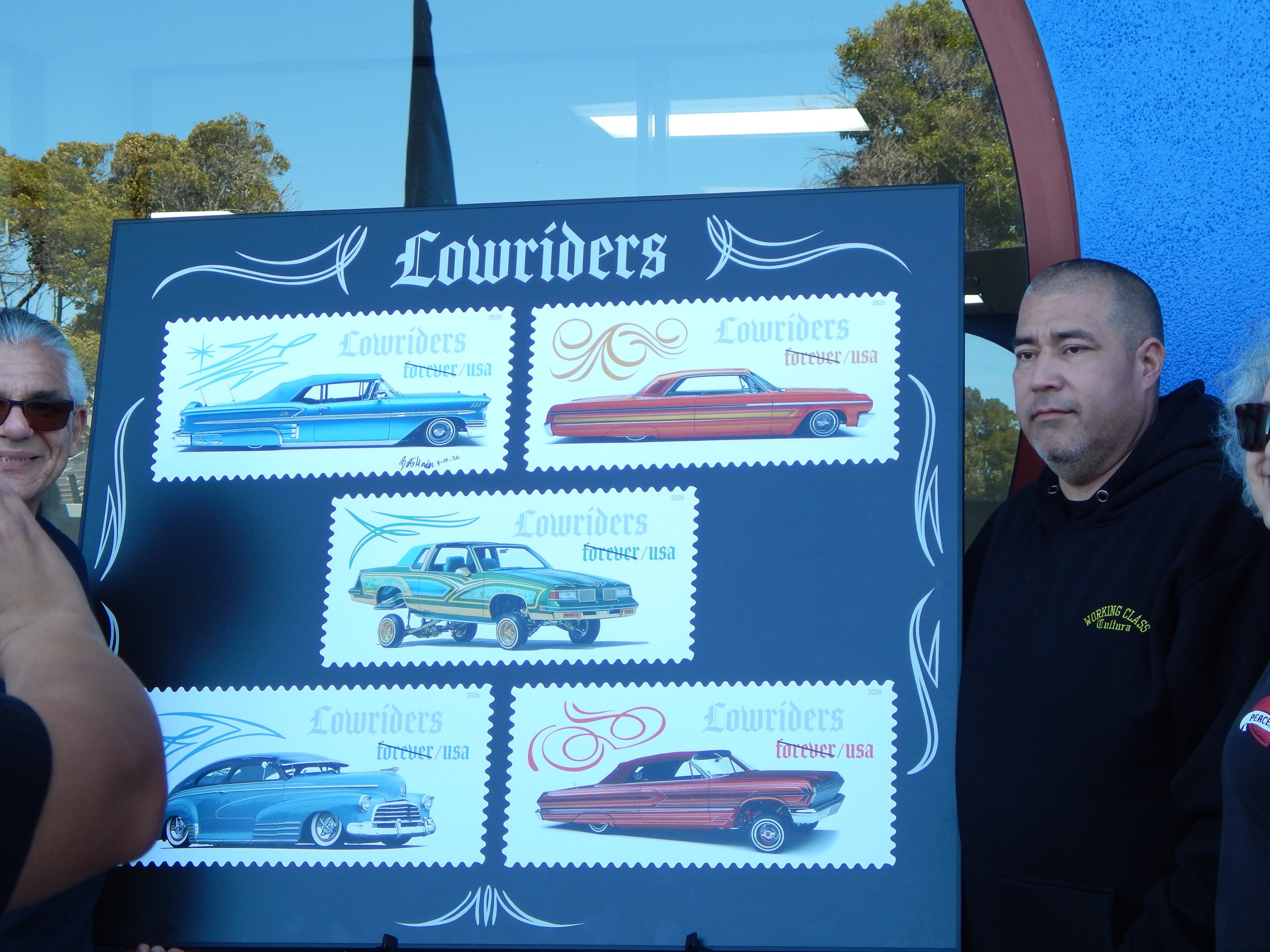 A group of people stands next to a display board showing five illustrated "Lowriders" postage stamps featuring classic cars.