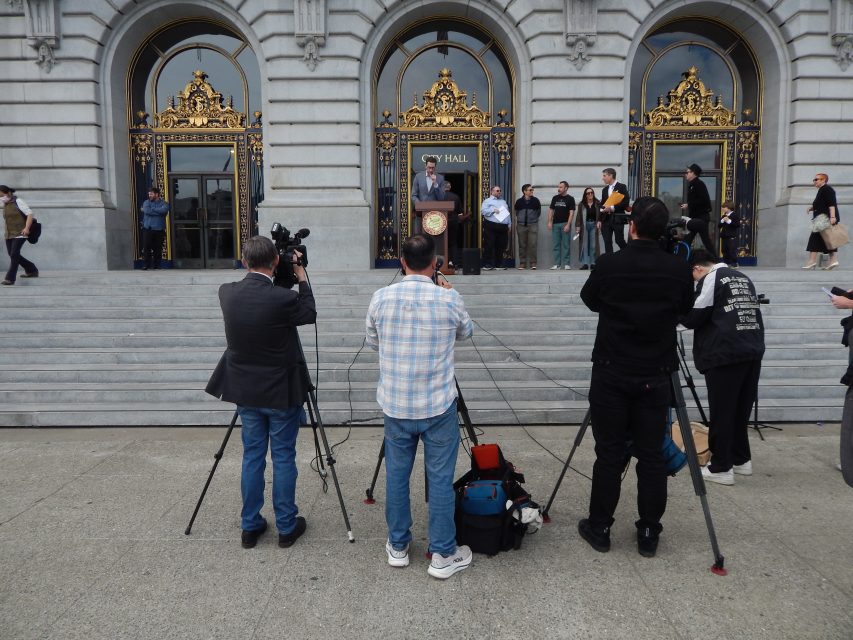 A person speaks at a podium on the steps of a large building while photographers and videographers capture the event. Several people stand near the entrance behind the speaker.