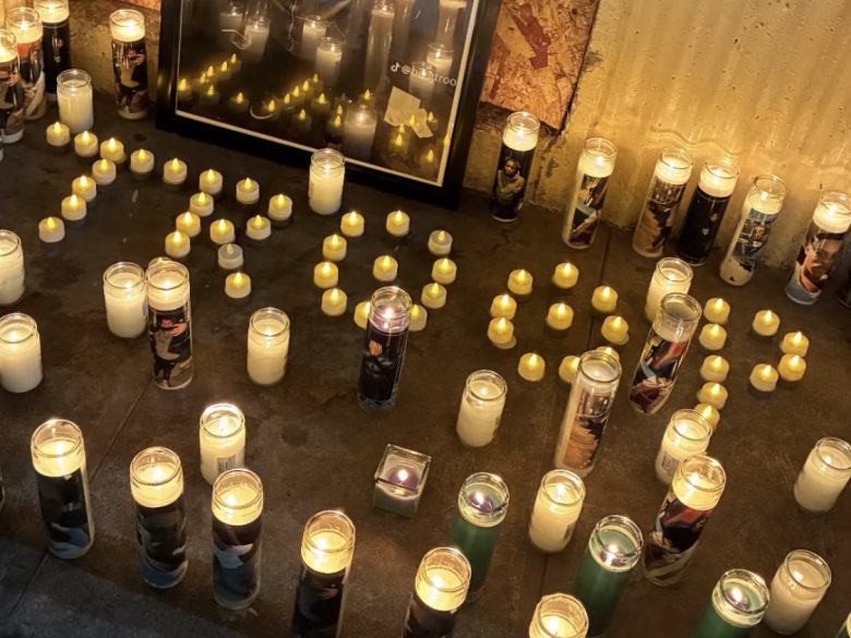 Dozens of lit candles arranged on a concrete surface form words, with more candles and a framed photo in the background, creating a memorial display.