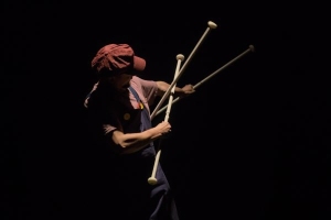 A person in a red cap and dark clothing performs on stage, dramatically crossing two crutches under a spotlight against a dark background.