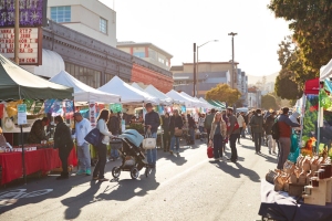 People walk and shop at an outdoor street market lined with tents and stalls on a sunny day.