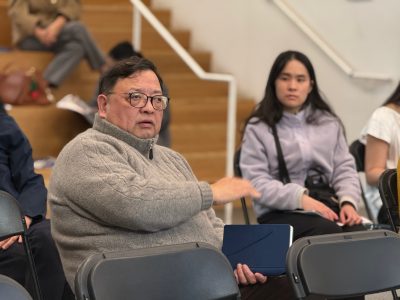 A man wearing glasses and a gray sweater sits and gestures with his hand while others sit nearby in a room with wooden steps.