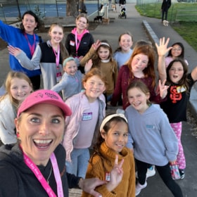 A group of girls and two adult women smile and wave at the camera outdoors on a paved path, some wearing name tags and lanyards.