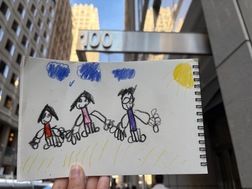 A hand holds a child's drawing of three people under blue sky and a yellow sun, with a city street and a building number 100 in the background.